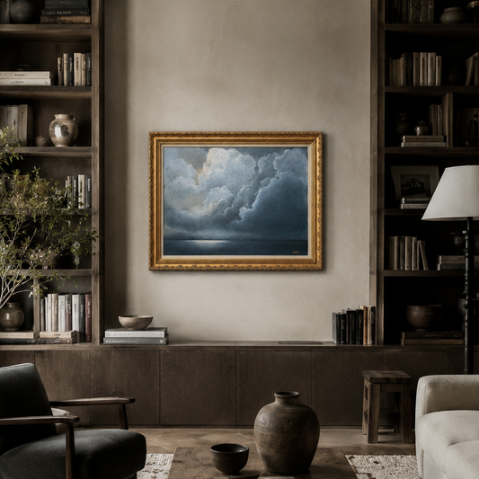 A framed landscape painting of stormy clouds hangs on a beige wall in a styled living room with dark wood bookshelves, a black armchair, a rustic coffee table, and neutral-toned furniture.