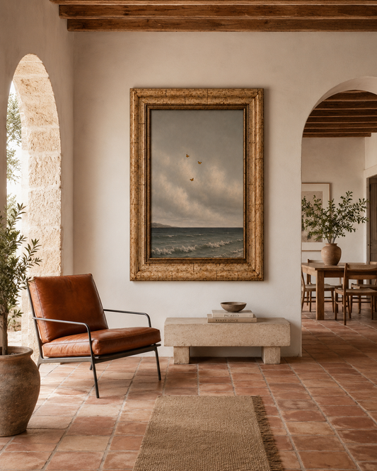 A large framed landscape painting of a stormy sea under a cloudy sky, displayed on a warm beige wall in a rustic, elegant interior with terracotta tile floors, a brown leather armchair, and wooden arches.
