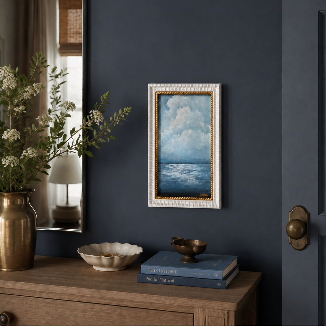 A framed vertical landscape painting of a blue ocean under a cloudy sky, styled on a dark navy blue wall above a wooden console table with books and a vase.