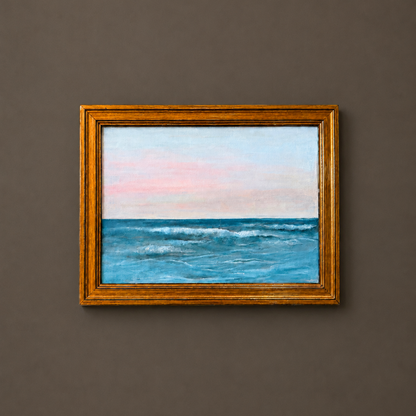 A framed painting of a blue ocean under a soft pink and light blue sky, set in a gold frame and mounted on a dark gray wall.