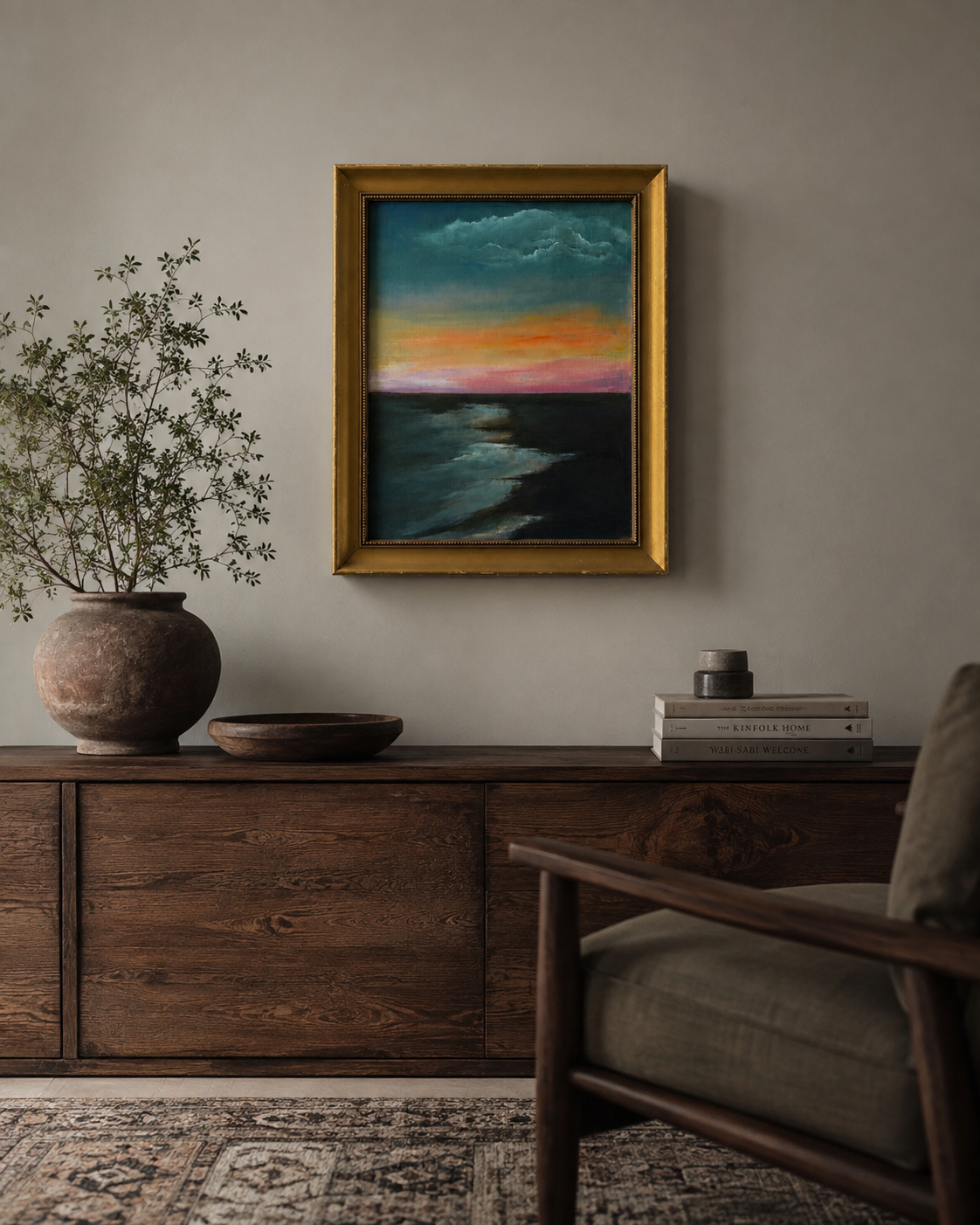 A framed landscape painting of a colorful sunset over the ocean hangs on a neutral wall above a dark wood console table in a styled living room setting.