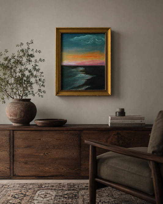 A framed landscape painting of a colorful sunset over the ocean hangs on a neutral wall above a dark wood console table in a styled living room setting.