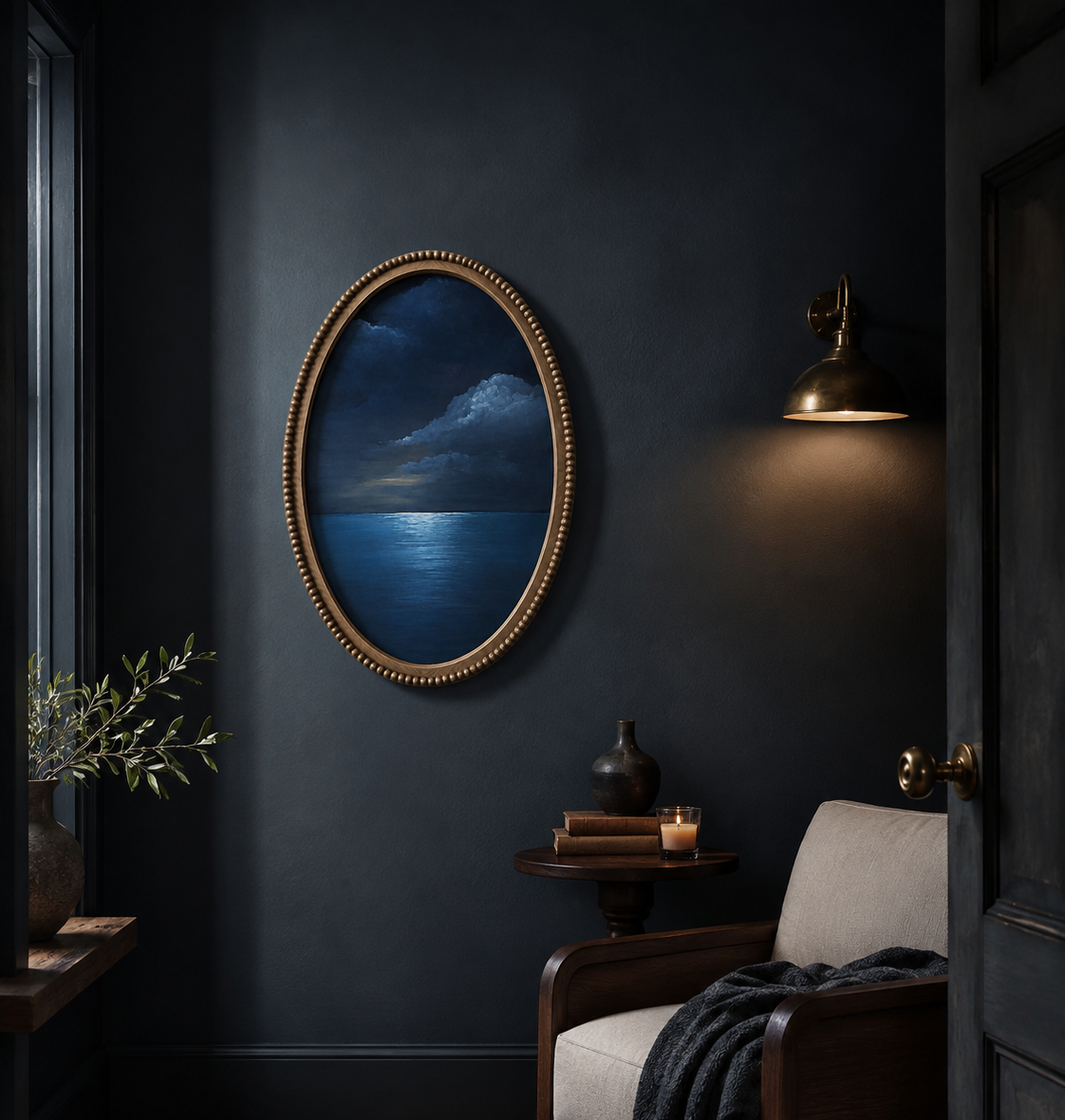 An oval framed wall art print depicting a dark blue night seascape with a stormy sky, styled in a moody, dark navy room next to a wooden armchair with a gray throw blanket.
