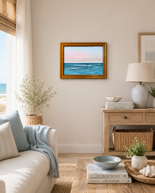 A framed square oil painting of a blue ocean under a pink and blue sunset sky, displayed on a beige wall in a beautifully styled coastal-themed living room.