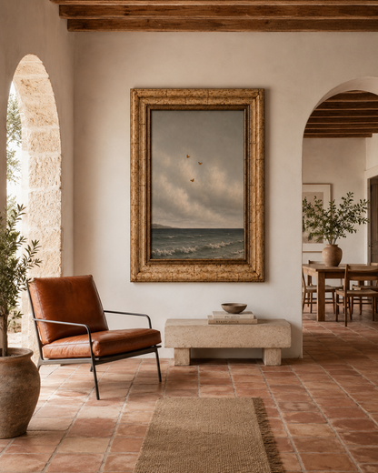 A large framed landscape painting of a stormy sea under a cloudy sky, displayed on a warm beige wall in a rustic, elegant interior with terracotta tile floors, a brown leather armchair, and wooden arches.