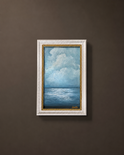 A framed vertical painting of a blue ocean under a cloudy sky, displayed on a dark brown wall. The painting features a textured white frame with a gold inner border.