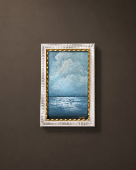 A framed vertical painting of a blue ocean under a cloudy sky, displayed on a dark brown wall. The painting features a textured white frame with a gold inner border.
